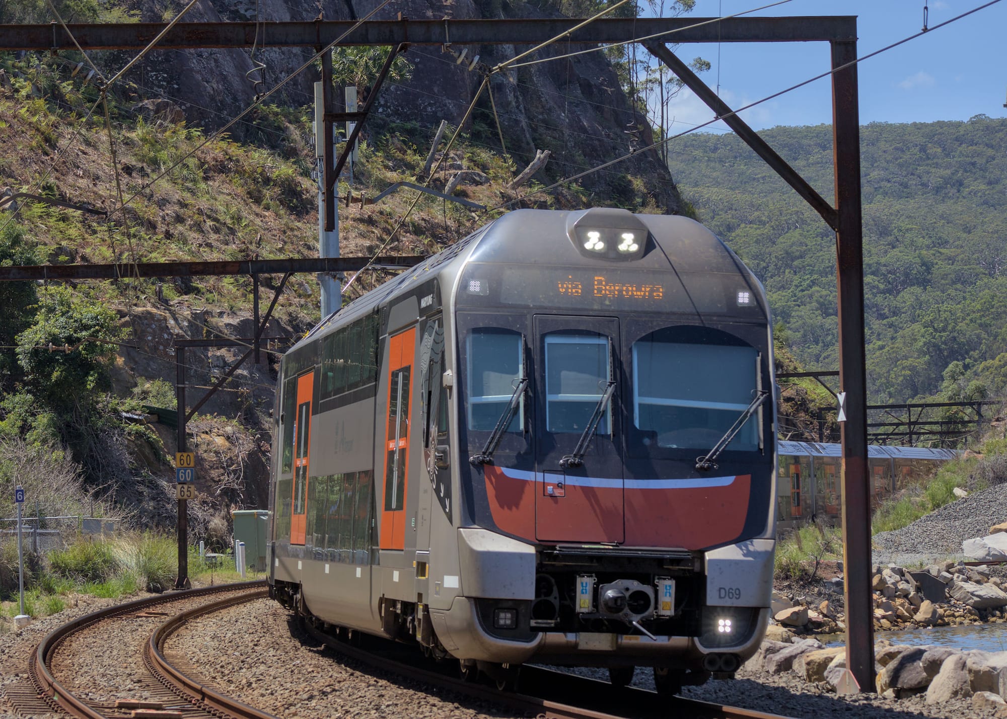 Silver, black and red Mariyung train rounds a curve between rocky bushland and Mullet Creek under blue skies