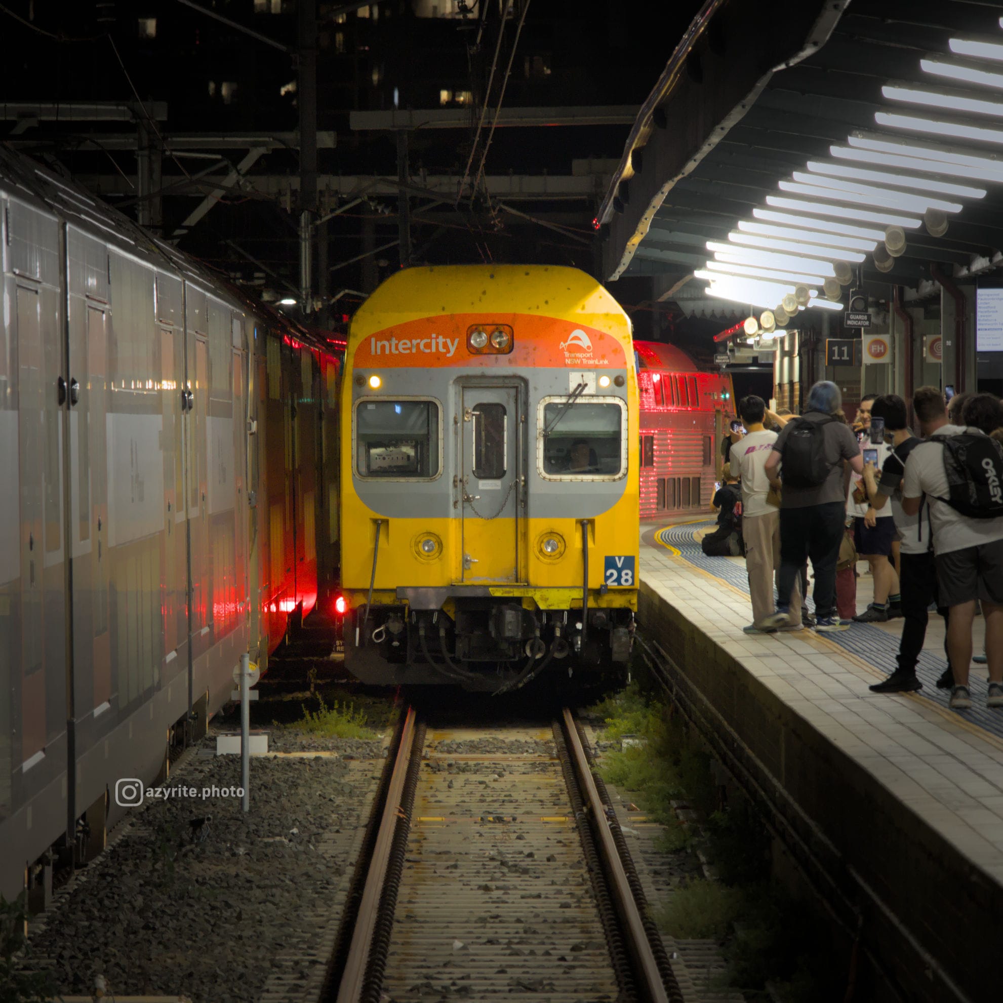 Head-on photo of V28 in its yellow, red and grey Intercity livery pulling into the brightly-lit Platform 11 at night where enthusiasts can be seen gathering