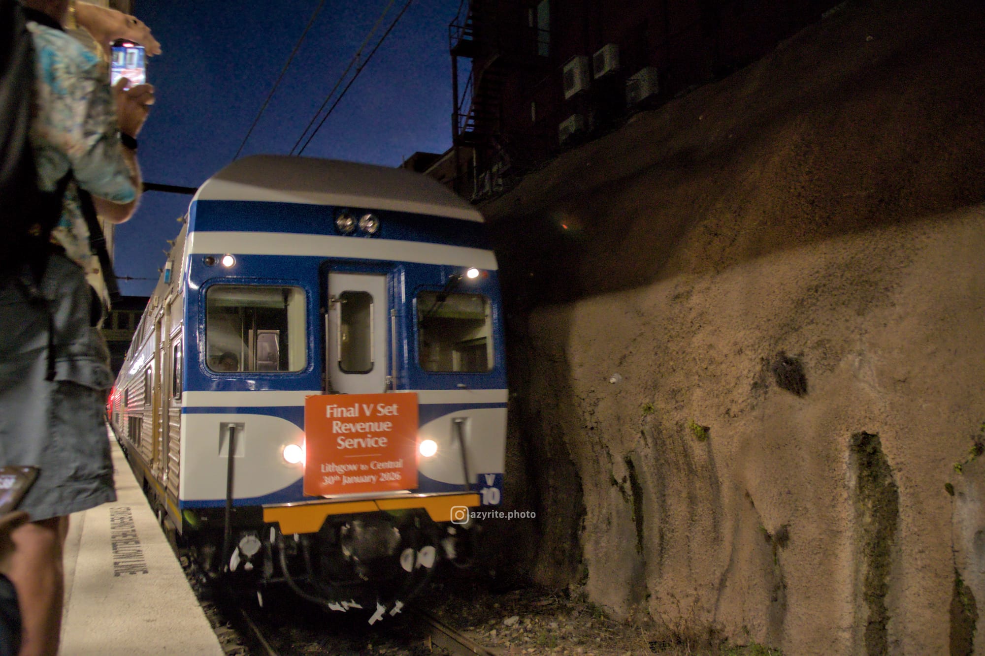 V10 with its original royal blue & white liveried front arrives into the platform before dawn with an orange headboard reading "Final V set Revenue Service" and "Lithgow to Central 30th January 2026"; a photographer with a phone is partially visible on the platform