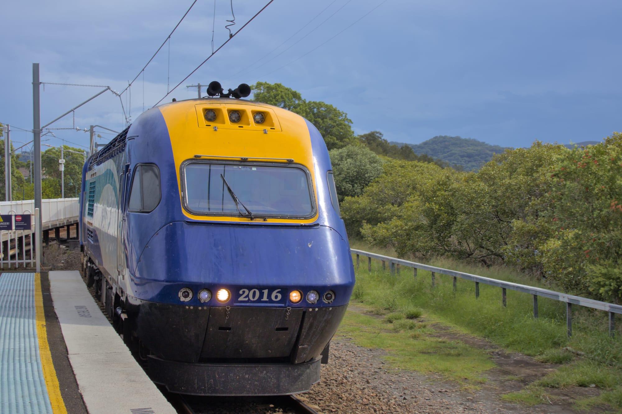 Blue and yellow power car running through a slightly curved platform, under overcast skies, green hills and trees are visible in the background