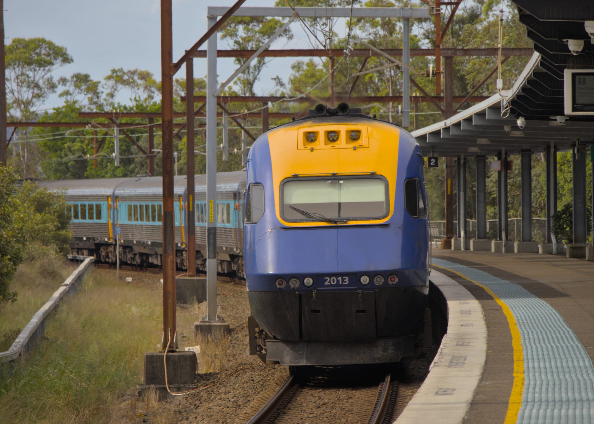 Blue and yellow power car at the end of the train beside a curved platform with yellow and blue tactile indicators, framed by rusted gantries and bush