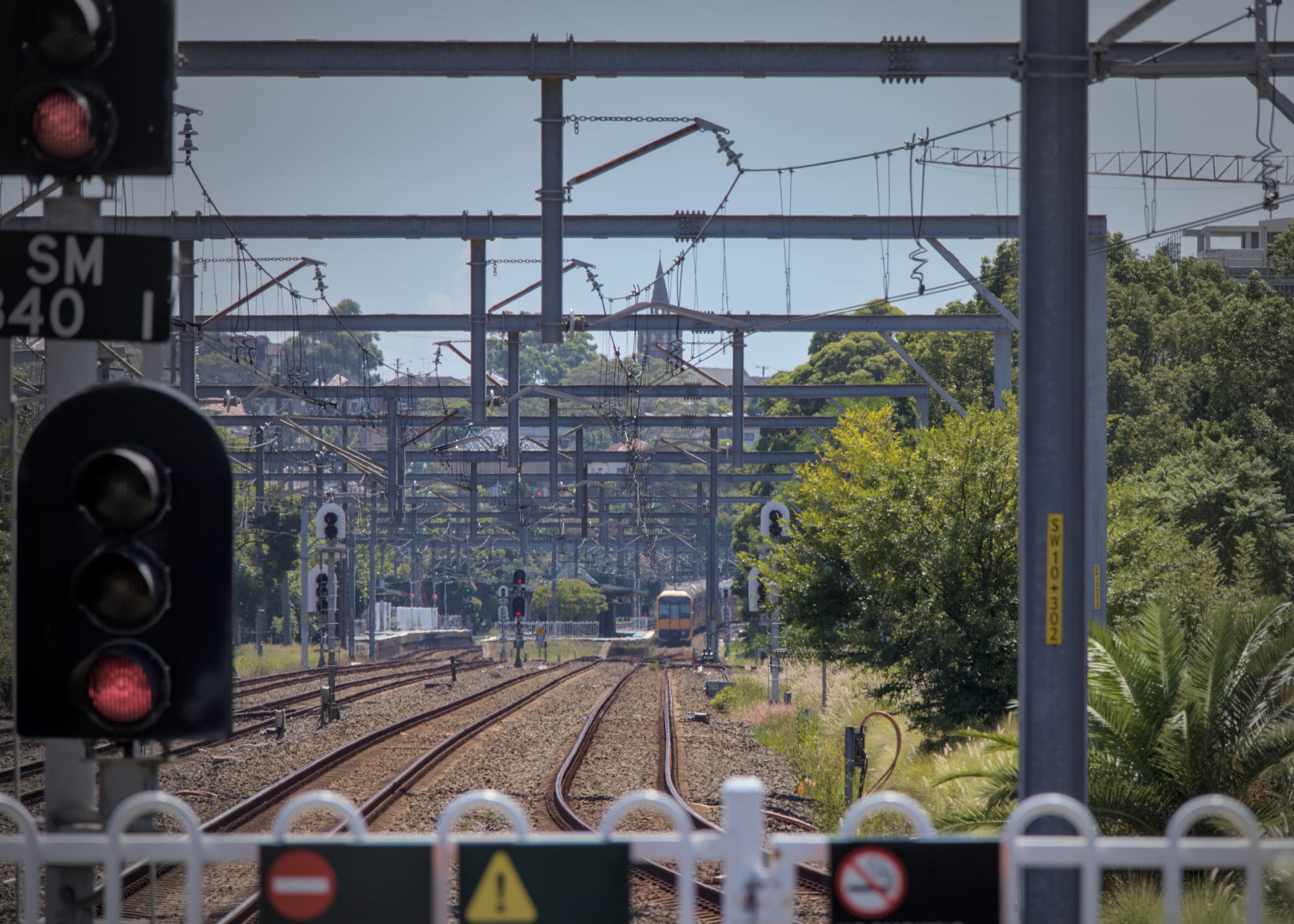 View up the line from the end of the platform, showing red signal lights in the foreground and multiple tracks extending under gantries with a distant Tangara at the next station