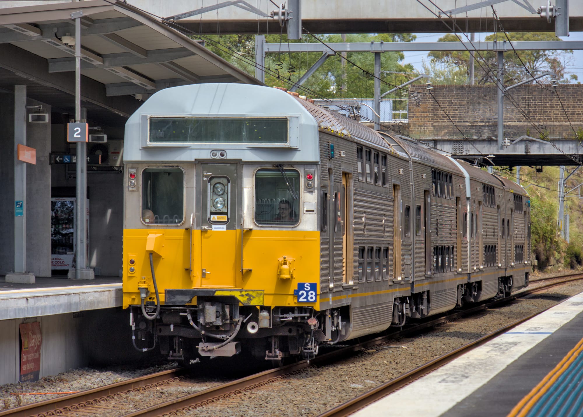 Rear and right side of silver S28 visible passing through Platform 2 at Jannali station under wires