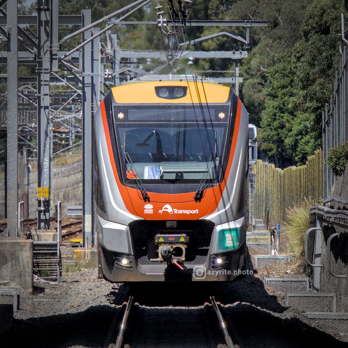 Front view of a modern silver orange and yellow train in daylight about to cross into the shadow of a bridge, emerging from a cutting
