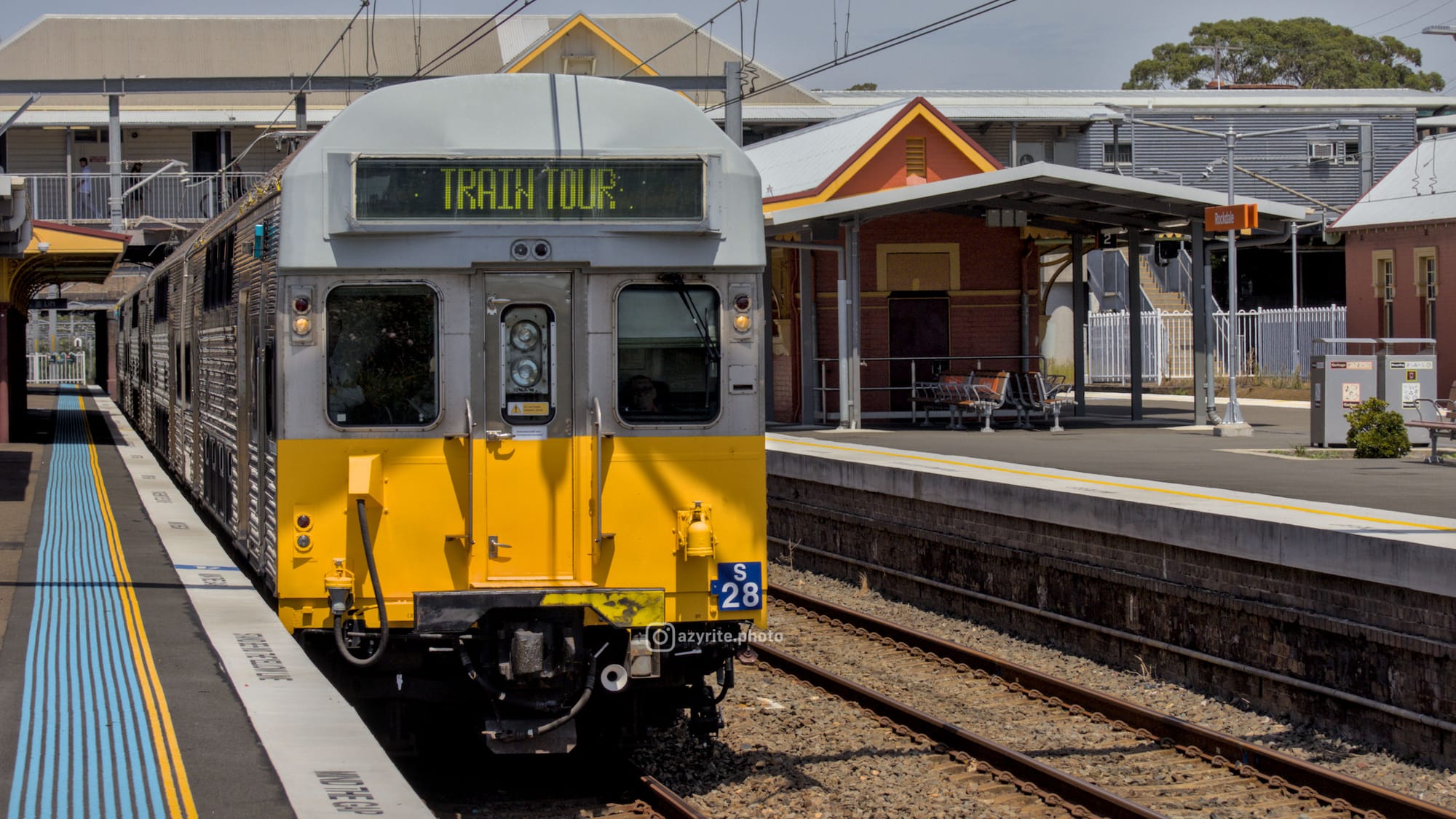 Silver and yellow heritage suburban train with "TRAIN TOUR" displayed on its flip-dot destination board passes a platform under overcast skies