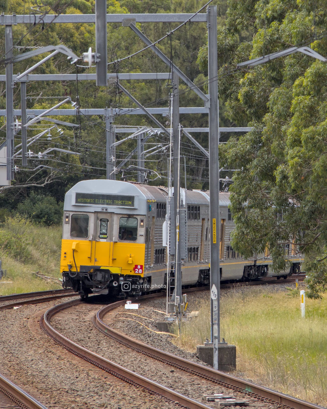 Silver and yellow train waiting on a curve before a raised white trainstop in amid a leafy scene framed by overhead wires and gantries