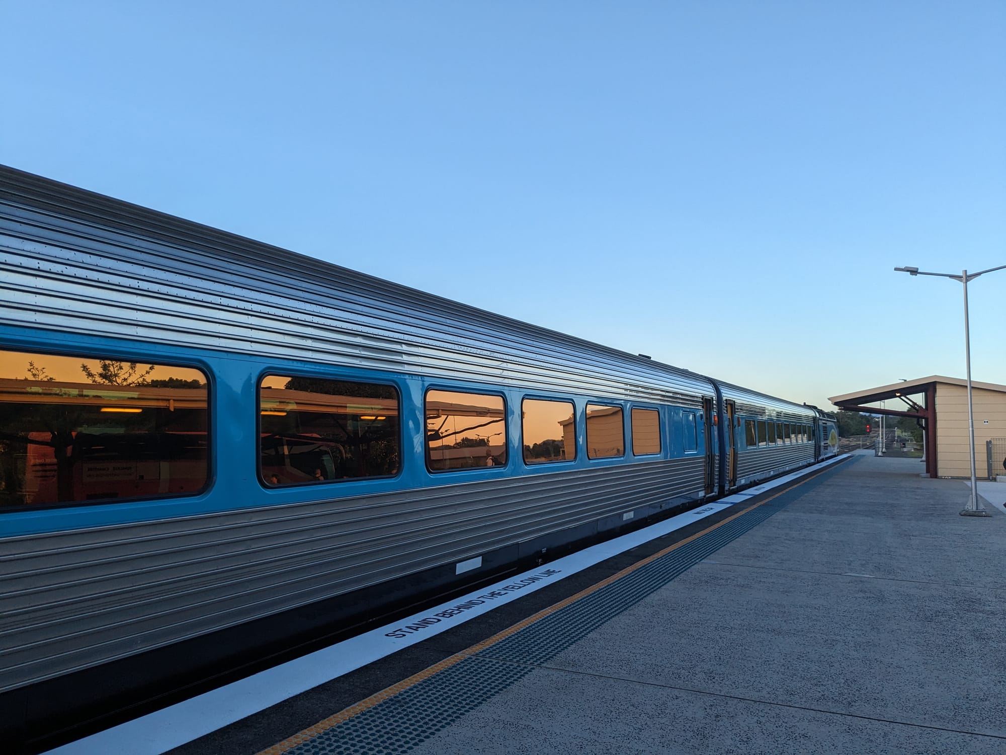 First two carriages and power car of a silver and blue passenger train stopped at an empty platform at dusk, with golden reflections in the tinted windows and a clear sky above