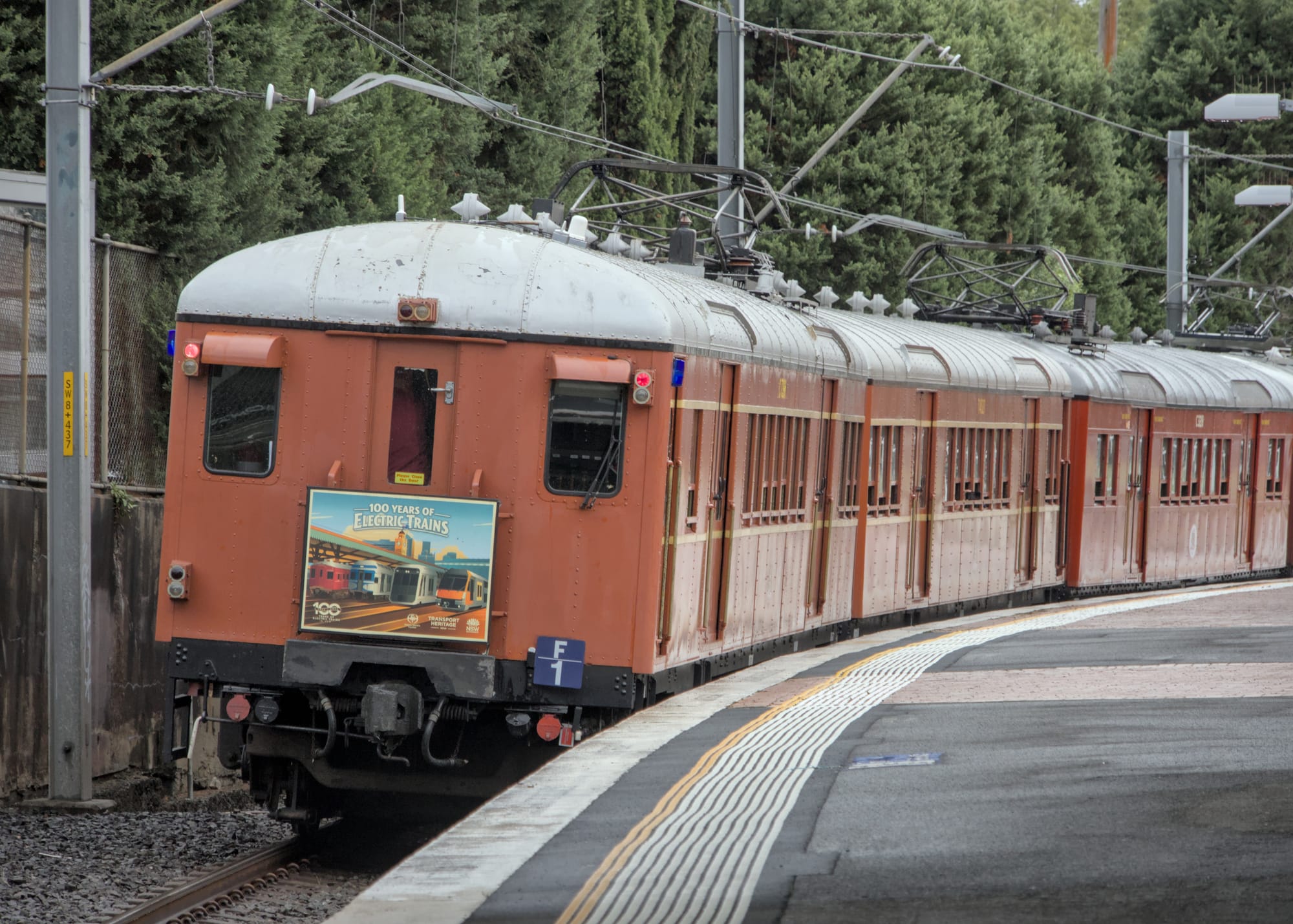 Rear view of the train in the previous image now rounding the curve past a tree-lined platform. The identical headboard is also affixed to the rear of the train.