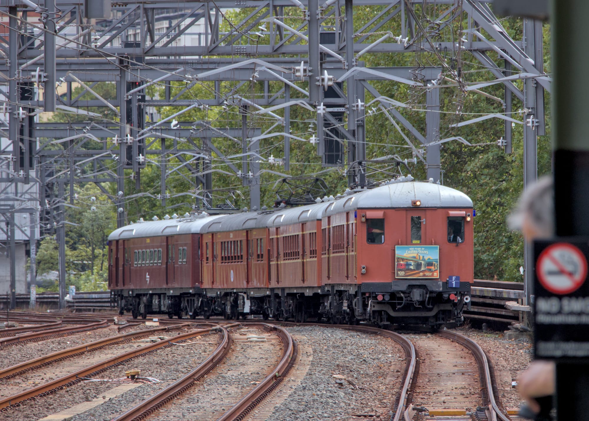 Front and left side view of silver-roofed F1 approaching at Central, under multiple steel gantries and wires. A green/black pole with a no smoking sign is visible in the foreground to the far right of the image but is out of focus.