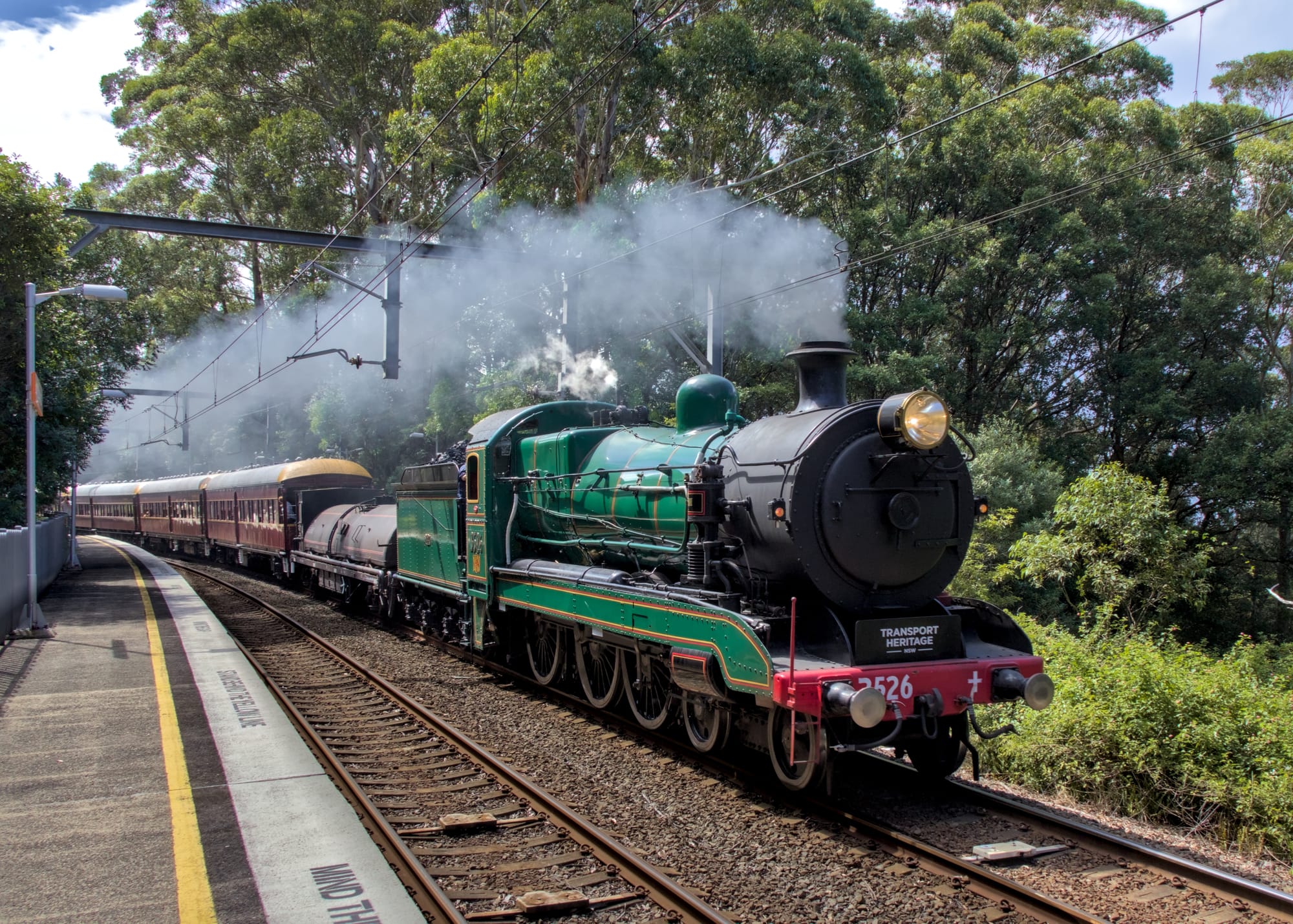 Front and side view of the shiny green and black steam locomotive with its tender in matching green, hauling its cream and maroon carriages around a slight curve at the station, light grey drifting from the smokestack. Lighting is very bright daylight. 