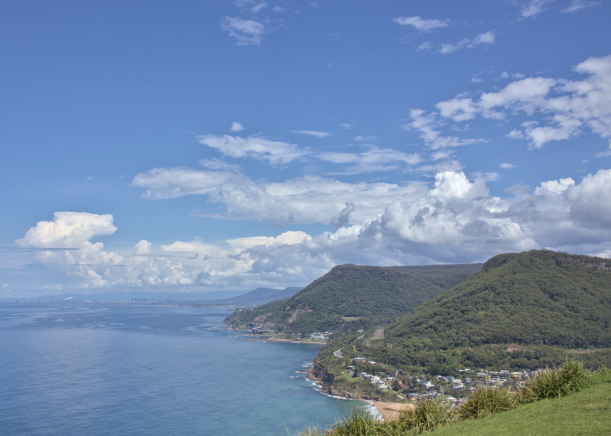 Panoramic coastal view with the Illawarra Escarpment visible on the right of the image, under a large blue sky with defined white clouds. Houses are visible at the foot of the hill closest.