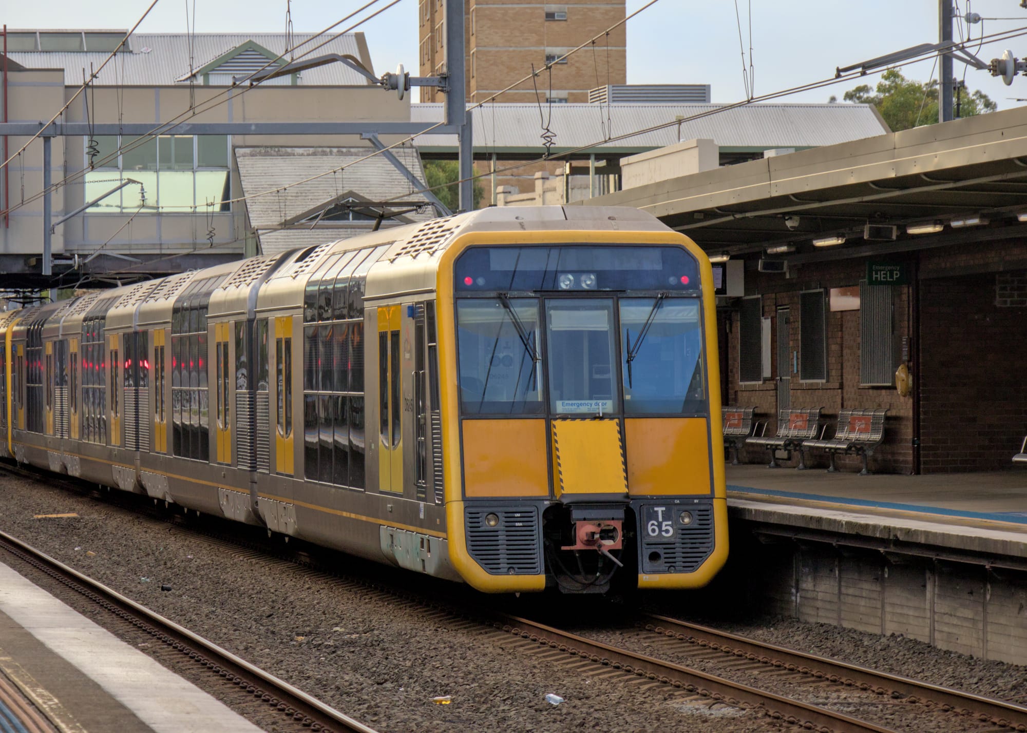 Yellow-fronted double-deck suburban train under wires at a platform with a footbridge visible in the background