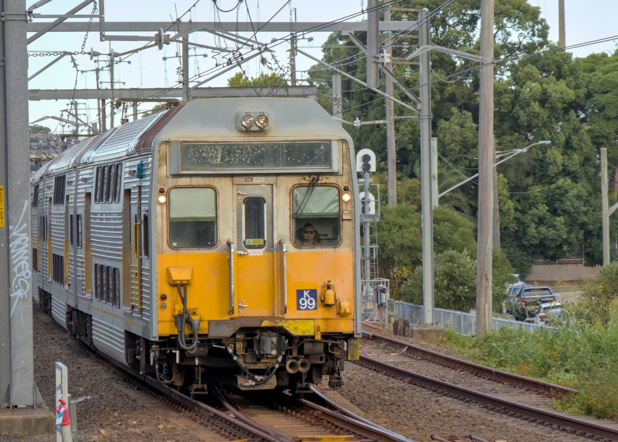 Front of a silver double-deck suburban train approaching, beneath overhead wires