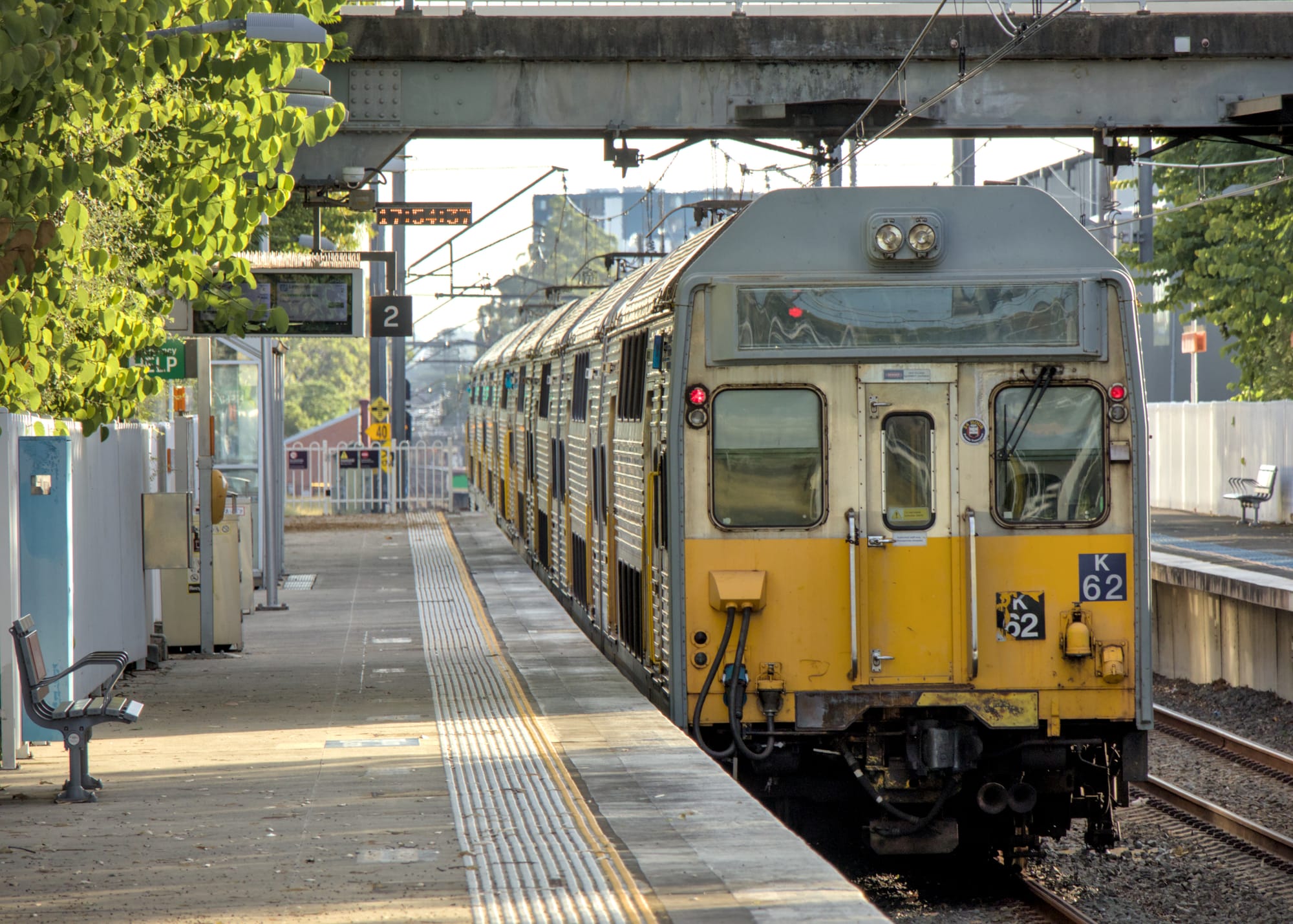 Rear view of silver suburban train leaving the platform, framed by an overpass and trees