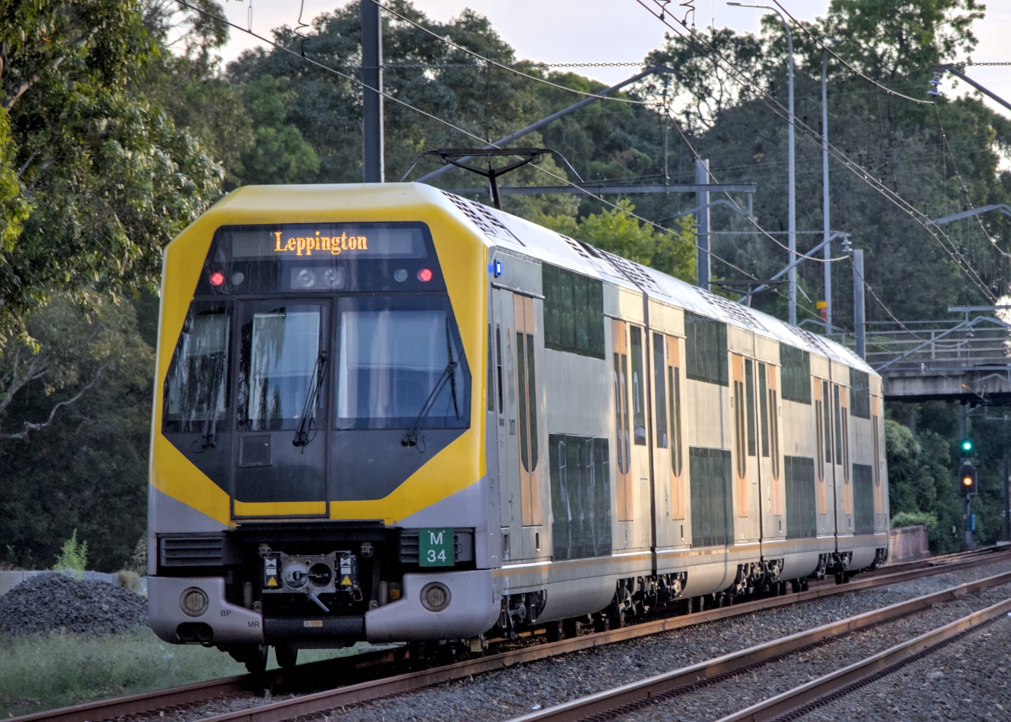 Modern yellow-fronted double-decker train moving along a tree-lined rail corridor