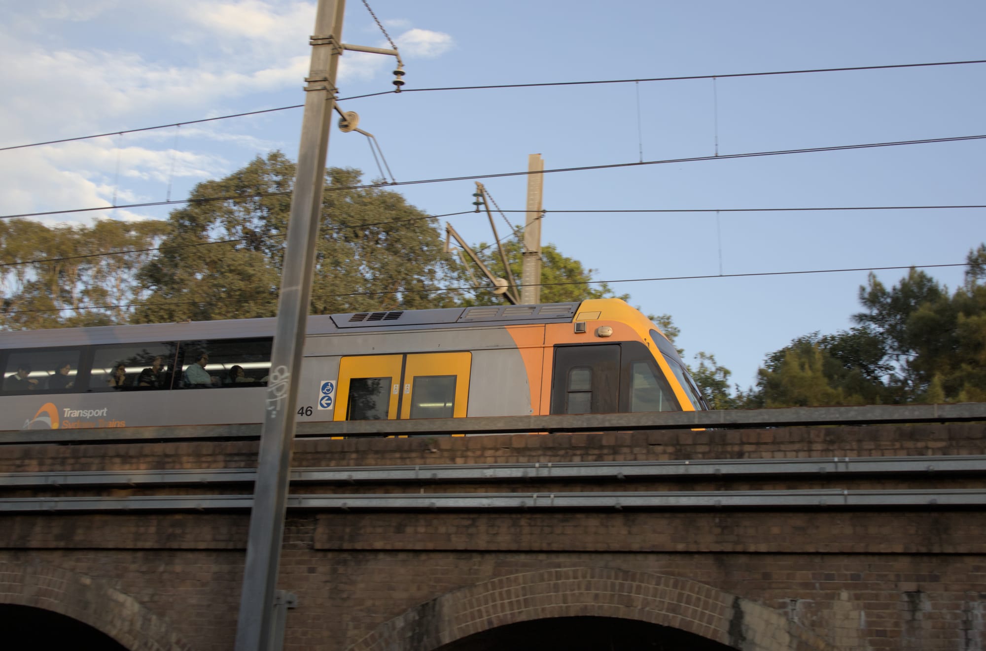 Low-angle side view of a silver and orange suburban train passing over a brick railway bridge