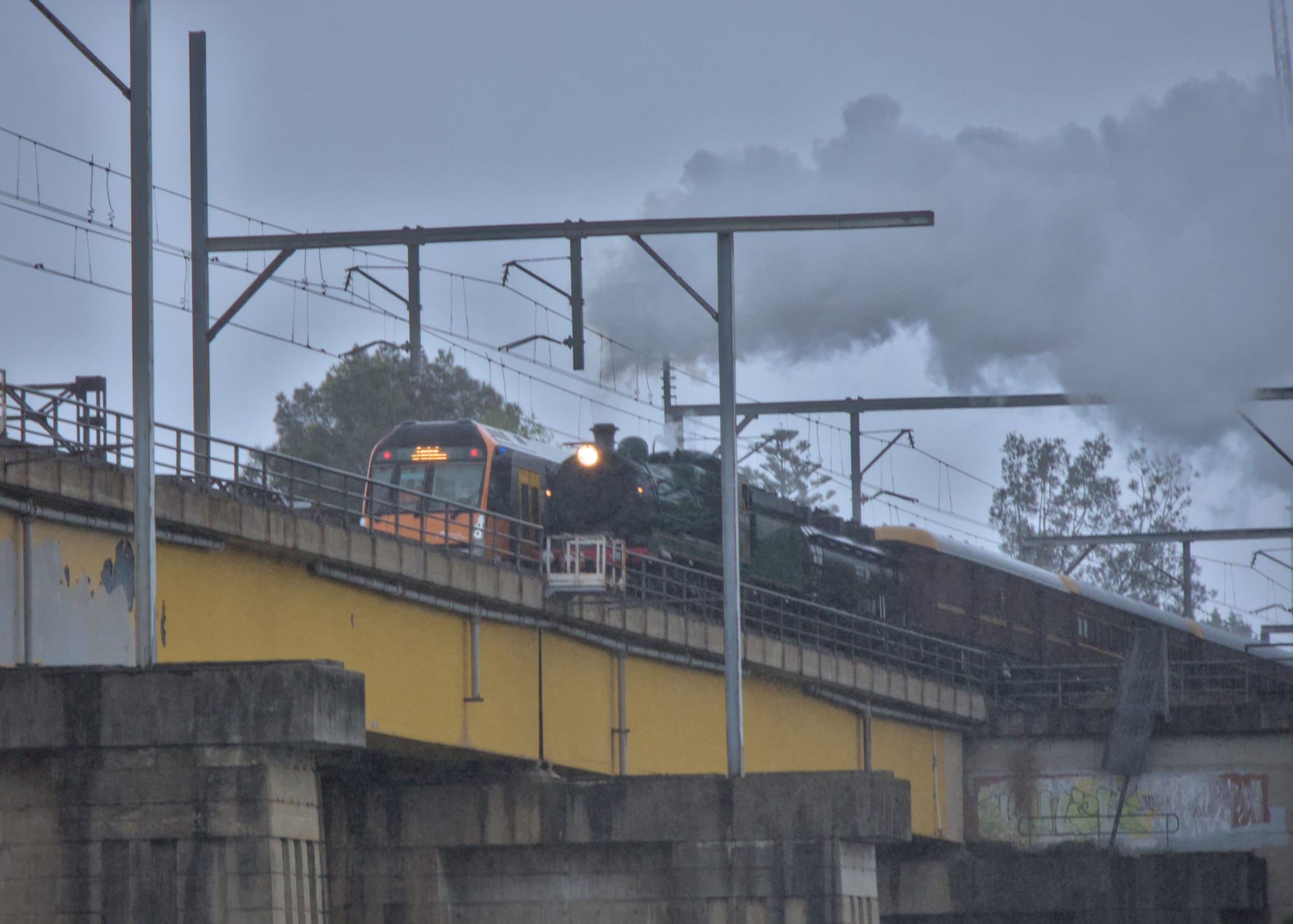 Steam train and modern passenger train passing each other on an elevated bridge in the wet