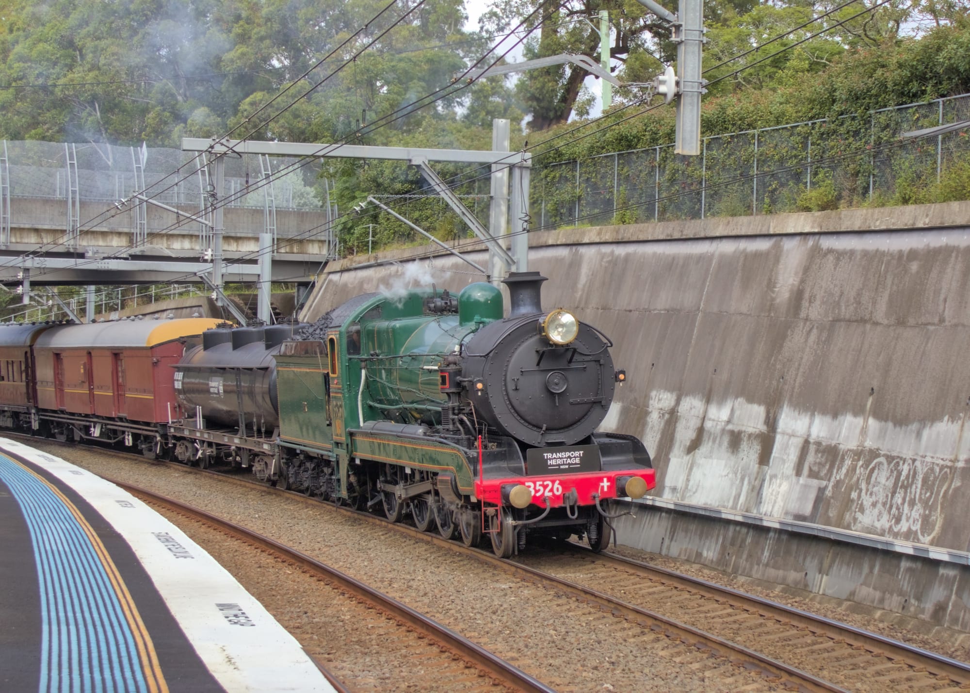 Partial side view of 3526 pulling carriages beside the station, next to a concrete wall and under an overpass