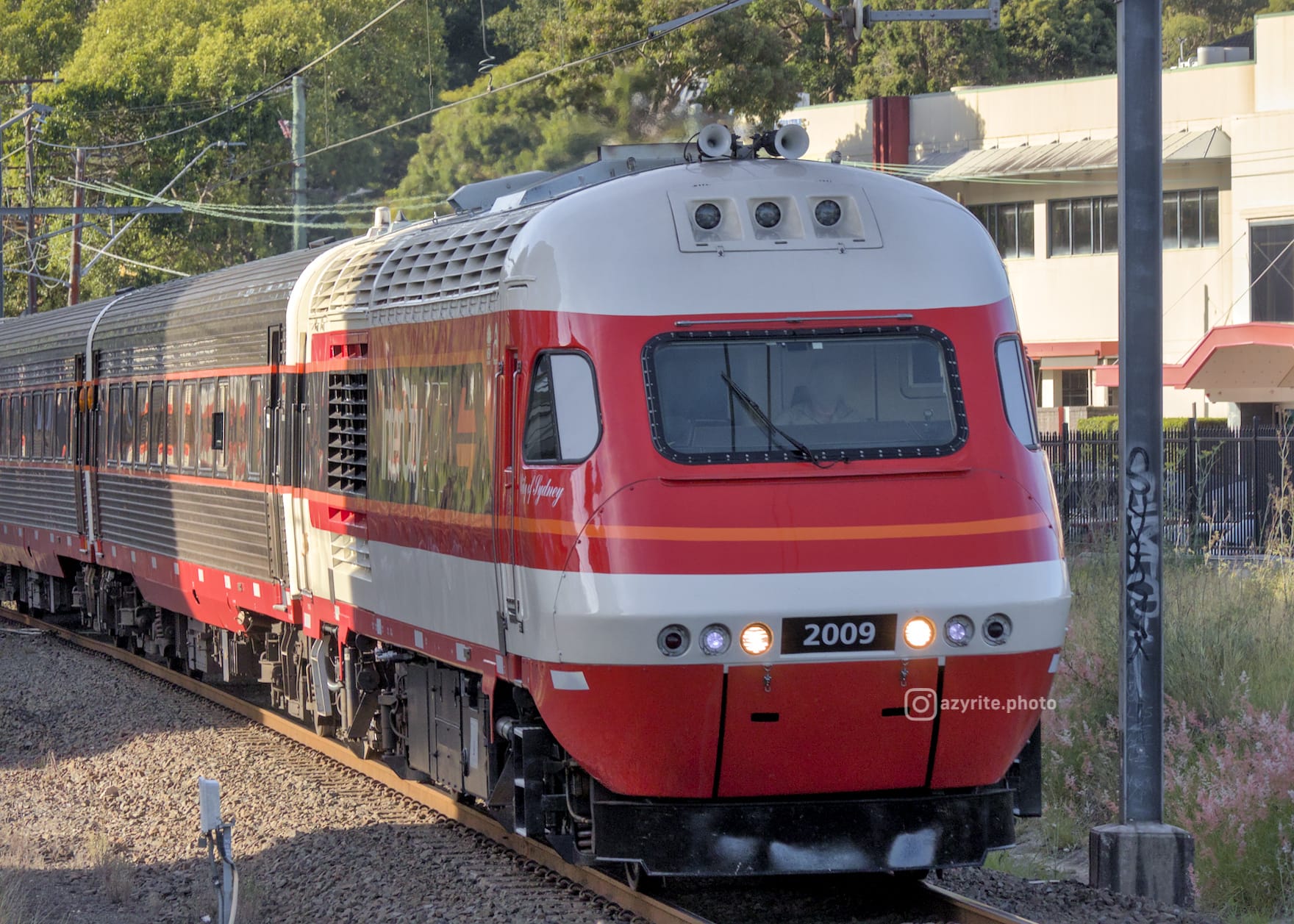 Red and white candy-liveried XPT power car leads steel carriages with leafy trees in the background