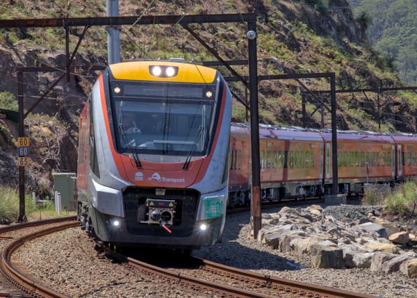 Silver and red liveried R set rounds a curve, framed by rusted gantries on a sunny day with forest hills in the background 