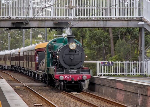 Feature image: Front view of a green and black steam locomotive pulling a train of heritage maroon and cream coloured carriages passing under a footbridge