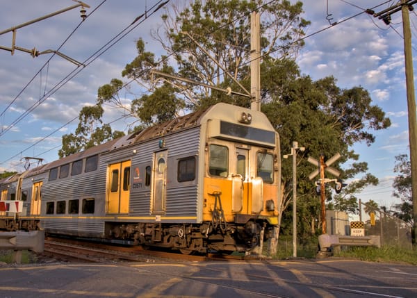 Silver double-decker suburban train crossing a road level crossing at sunset, during golden hour, framed by overhead power lines and gum trees in the background.