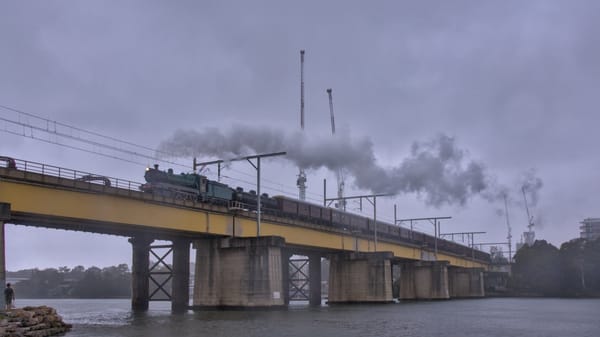 Green steam locomotive leading a long train of heritage locomotives over a long yellow steel beam bridge on a gloomy day